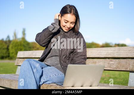 Young brunette girl sits in park with laptop, watches video or browses internet, rests outdoors on bench in sunny park Stock Photo