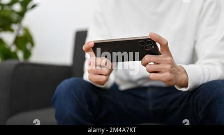 A young man sits on sofa playing a mobile game, watching movie or football match on his smartphone. cropped image Stock Photo