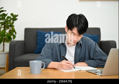 Focused young Asian male college student writing diary or taking notes on a school book, doing homework in his living room. Stock Photo