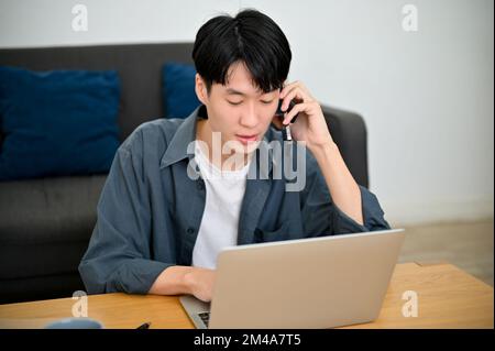 Handsome and focused young Asian man talking on the phone while using laptop computer and working in his living room. Stock Photo