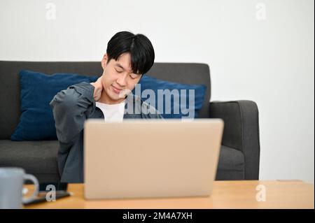 Tired young Asian male freelancer sits in the living room touching his neck, getting neck pain, suffering from office syndrome, remote working at home Stock Photo