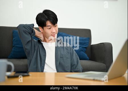 Tired young Asian male freelancer sits in the living room using laptop computer, touching his neck, getting neck pain, remote working at home. Stock Photo