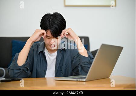 Thoughtful and stressed young Asian male college student pensively thinking about his school project, sitting in the living room. Stock Photo