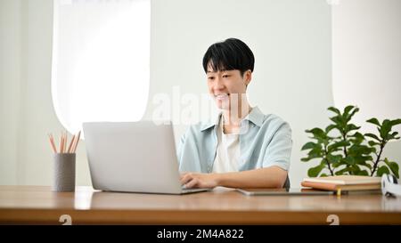 Happy and handsome young Asian man working on his tasks on laptop in his minimal office. working, managing project, doing online homework. Stock Photo