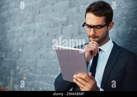 Giving it some thought. a handsome young businessman using his tablet while standing against a grey facebrick wall. Stock Photo