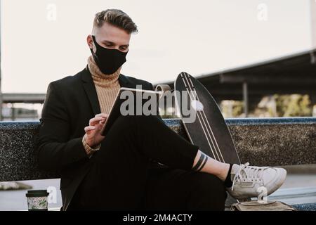 Skater with facial mask using a tablet sitting outdoors Stock Photo