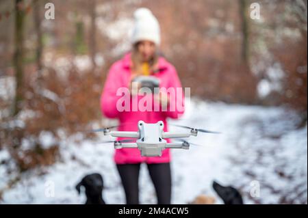 Young woman in pink winter jacket standing in snowy forest using a controller to direct and manage a drone floating in front of her. Focus to the dron Stock Photo