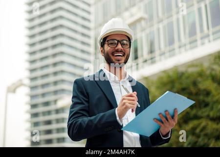 Indian construction architect in suit and hardhat making notes against modern building Stock Photo