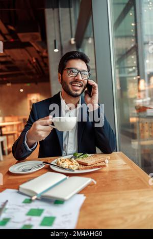 Happy indian businessman is talking phone with client during lunch time in cozy cafe Stock Photo