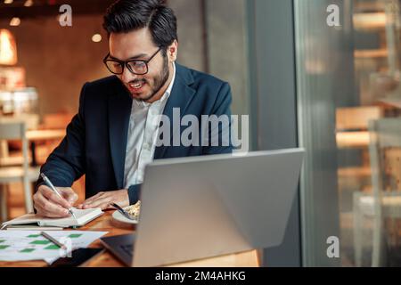 Indian businessman is making notes in notepad during working day in cafe. Blurred background Stock Photo