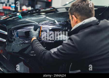 Rear view of man squatting by black car to apply PPF paint protection film on headlight in a garage. Indoor horizontal shot. High quality photo Stock Photo