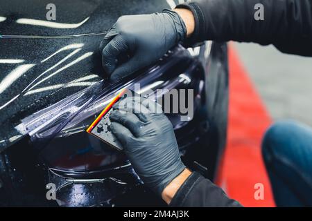 Close up of person wearing black gloves applying PPF paint protection film to headlight of car in a garage. Car detailing. Horizontal indoor shot. High quality photo Stock Photo