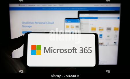 Person holding mobile phone with logo of productivity software Microsoft 365 on screen in front of business web page. Focus on phone display. Stock Photo