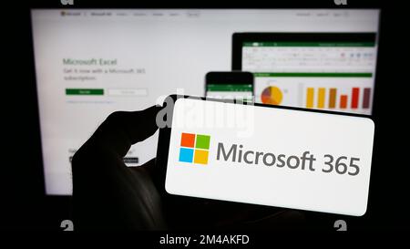 Person holding cellphone with logo of productivity software Microsoft 365 on screen in front of company web page. Focus on phone display. Stock Photo
