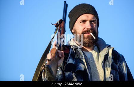Man brutal bearded guy gamekeeper blue sky background. Brutality and masculinity. Hunter with rifle gun close up. Guy bearded hunter spend leisure Stock Photo