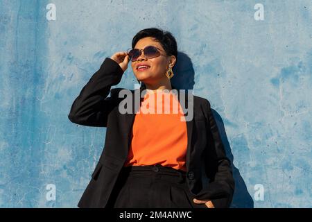 From below confident adult ethnic female entrepreneur with short dark hair in stylish suit and sunglasses smiling and looking away while standing agai Stock Photo