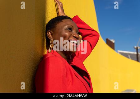 Side view of smiling young stylish adult African American female with short dark hair in stylish suit and earrings looking away standing near yellow w Stock Photo