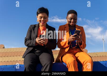Positive multiracial female friends in stylish suits and earrings sitting on blue fence and texting message on smartphones Stock Photo