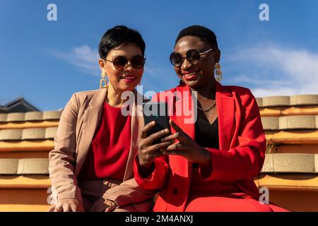 Cheerful adult African American woman with short dark hair in stylish outfit and sunglasses smiling while showing video on smartphone to ethnic female Stock Photo