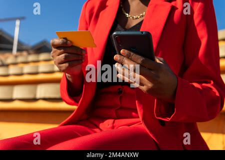 Cropped unrecognizable adult African American woman in stylish clothes with credit card making online purchase via smartphone while sitting on stairs Stock Photo