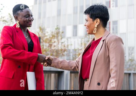 Positive adult multiracial female colleagues business partners with short hair in stylish suits smiling and looking at each other while shaking hands Stock Photo