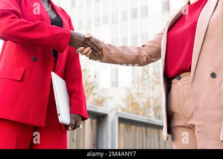 Cropped unrecognizable positive adult multiracial female colleagues business partners with short hair in stylish suits shaking hands standing on stree Stock Photo