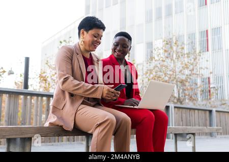 Stylish elegant adult multiracial female colleagues with short dark hair in stylish suits sitting in bench near modern building and using laptop and s Stock Photo