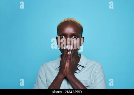 Astonished young African American female in stylish shirt with short blond hair and painted nails covering mouth in shock and looking at camera agains Stock Photo