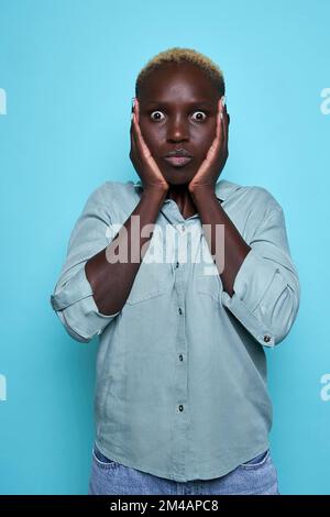 Astonished young black woman in shirt with curly blond hair touching cheeks and looking at camera with opened mouth against blue background Stock Photo