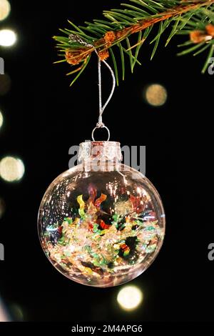 Closeup of transparent glass bauble with colorful confetti hanging on branch of Christmas tree in dark room with glowing lights Stock Photo