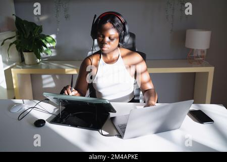 Young African American woman listening to music and drawing on graphic table with stylus while sitting at desk and working on remote freelance project Stock Photo