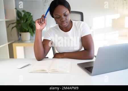 Young African American woman scratching head with pen and thinking over homework assignment while sitting at table with notepad and laptop and studyin Stock Photo