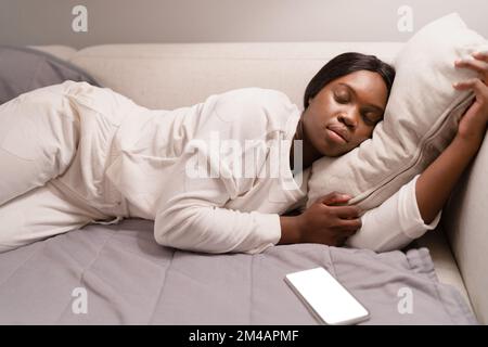 Young black woman in white sleepwear lying on soft pillow and sleeping on sofa near smartphone with blank screen in living room in evening at home Stock Photo