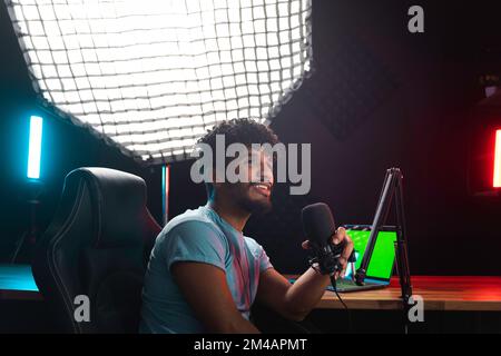 Happy young Hispanic male blogger in blue t shirt looking away and talking into microphone while sitting near laptop with chromakey screen and recordi Stock Photo