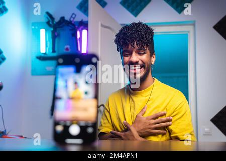 Cheerful young Hispanic blogger crossing hands on chest and smiling while sitting at desk behind smartphone and telling funny story to vlog audience i Stock Photo