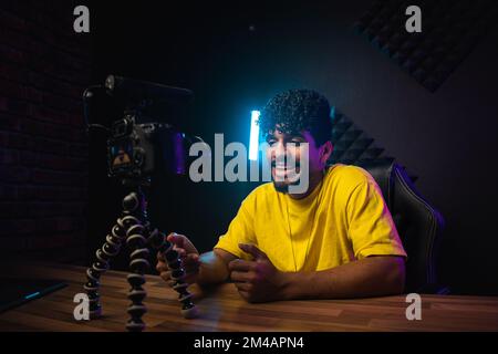 Optimistic young Hispanic male vlogger in yellow t shirt smiling and talking to audience while sitting at table in front of camera and recording video Stock Photo