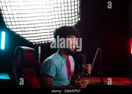 Optimistic young Hispanic male blogger with curly hair looking away with smile and speaking into microphone against lamp during podcast recording in o Stock Photo
