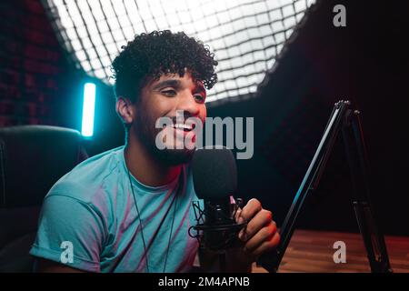 Optimistic young Hispanic male blogger with curly hair looking away with smile and speaking into microphone against lamp during podcast recording in o Stock Photo
