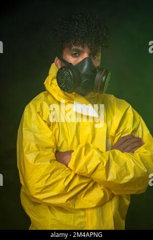 Young Hispanic man in yellow hazmat suit and respirator looking at camera while standing in smoke in dark illegal laboratory Stock Photo