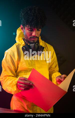 Young Hispanic man in yellow hazmat suit reading papers in folder while standing under neon light in obscure illegal laboratory at night Stock Photo