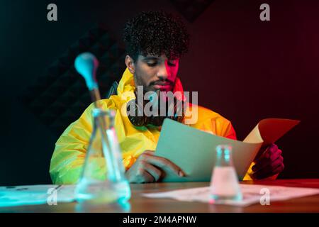 Glass vials with chemicals and pipette placed on table near man in hazmat suit reading file during work in obscure illegal laboratory Stock Photo