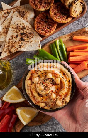 Top view of anonymous person putting bowl of yummy hummus near wood slab with slices of assorted vegetables and lemon and tray with tortilla chips and Stock Photo