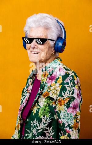 Happy senior female in floral blouse and pixel sunglasses looking at camera with smile while listening to music against yellow background Stock Photo