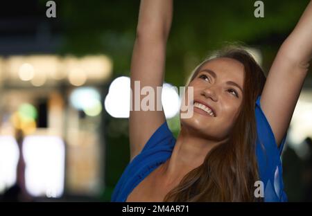 Happy young brunette looking up with smile and raising arms on blurred background of street at night Stock Photo
