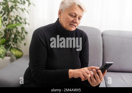 Happy mature old 60s woman holding smartphone using mobile phone app for video call, laughing while watching funny video, feeling excited winning Stock Photo