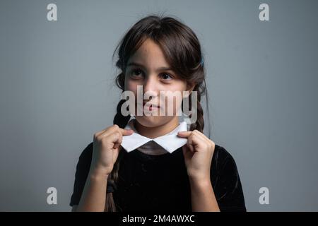 Portrait of little girl with Wednesday Addams costume during Halloween. Serious expression and dark atmosphere with dark background. Stock Photo