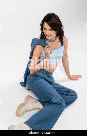happy brunette woman in pants and boots sitting with blue jacket and looking away on grey Stock Photo