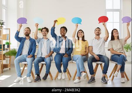 Positive diverse people sit in row and express their opinion by raising colored speech bubbles. Stock Photo
