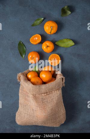 sweet tangerines with green leaves fall out of a crafting bag on a dark blue background. Dark photo. Top view Stock Photo