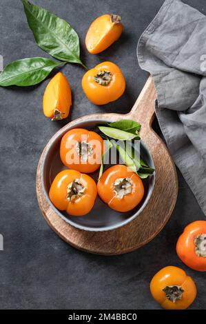 Fresh ripe persimmon fruits in bowl on a cutting board on a dark background with slices and leaves. The concept of healthy nutrition and vitamins. Top Stock Photo